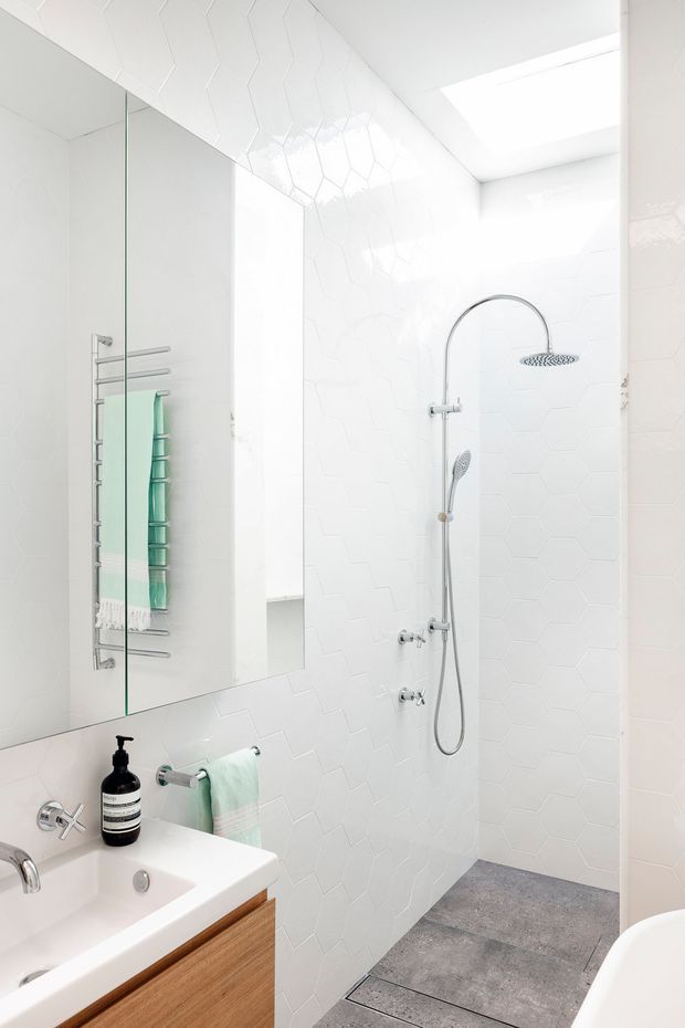 Bright contemporary bathroom with a walk-in shower, white geometric wall tiles, grey floor tiles, chrome fixtures, a large frameless mirror, skylight, white vanity and timber cabinetry. A freestanding bathtub is partially visible at the lower right.