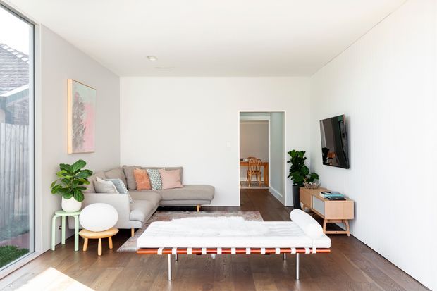 Bright, compact contemporary living room with white walls and ceiling, dark timber flooring, a light grey sectional sofa, wall-mounted television, and a low timber media console. A large rug and a distinctive low coffee table occupy the centre, while indoor plants and pastel cushions add softness. Full-height glazing on the left brings in abundant daylight, and an open doorway reveals a dining area beyond.