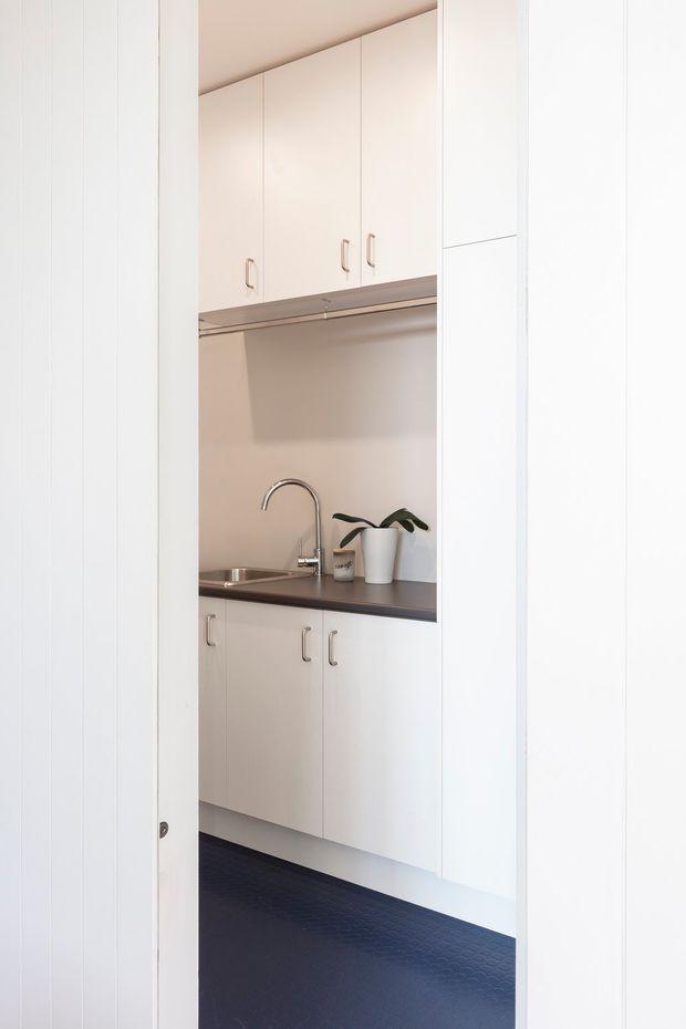 A compact built-in kitchenette or utility space viewed through a doorway, with white floor-to-ceiling cabinetry, upper cupboards, a dark benchtop, stainless-steel sink and curved tap. The room has white vertical wall panelling and a dark textured floor, creating a clean, simple and highly functional look.