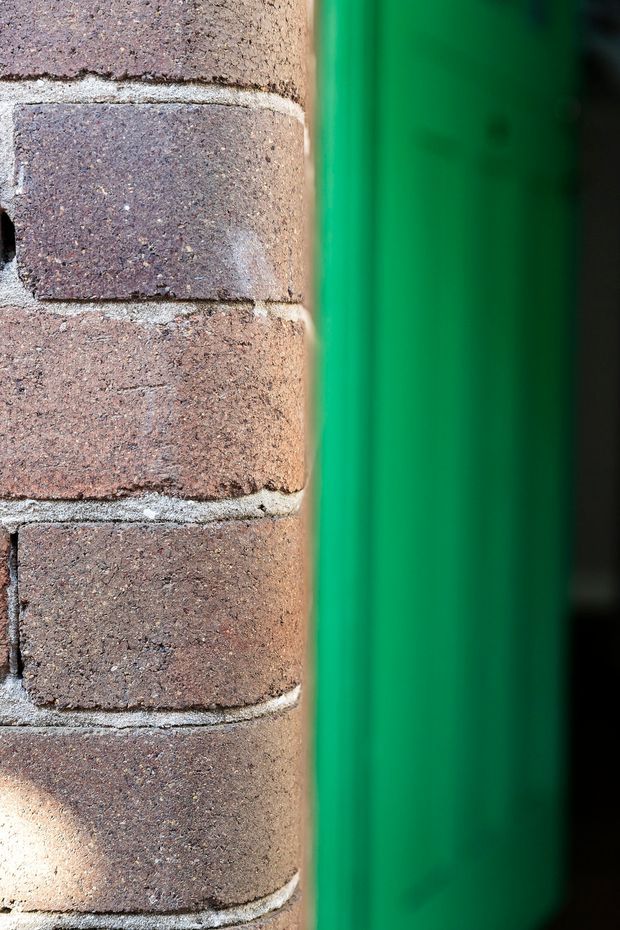 Close-up architectural detail showing rough reddish-brown brick masonry with pale mortar joints beside a vivid green vertical metal or painted frame/panel. The image is tightly cropped and abstract, with no identifiable room or broader building context.