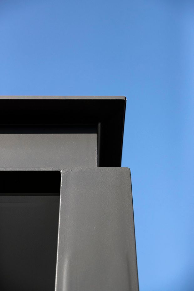 Close-up upward view of a sharply detailed modern building facade against a clear blue sky. The image emphasizes dark charcoal-grey exterior surfaces, deep recessed openings, crisp rectangular geometry, and a projecting flat roof or canopy with strong shadow lines.