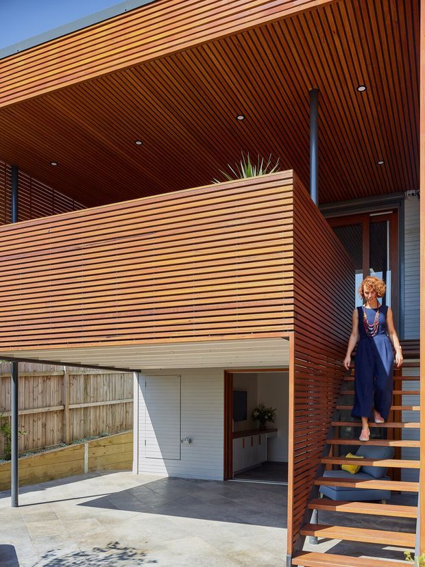 Exterior view of a contemporary multi-level home featuring extensive horizontal timber slat cladding and screening, a timber-lined covered balcony or entry canopy, open-riser timber stairs, dark metal posts, and a partially visible carport or ground-level entry. The facade combines warm timber with white horizontal siding and dark grey paving. A person is descending the stairs.