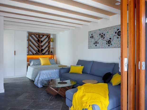 Compact open-plan living and sleeping space with a blue sectional sofa, yellow cushions and throw, and a bed positioned behind it. White walls and exposed white ceiling beams create a bright interior, while dark large-format floor tiles and warm timber-framed glazing add contrast. A built-in timber wine rack and colourful artwork are visible at the rear.