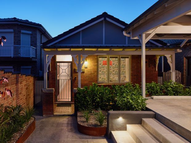 A dusk exterior view of a compact brick bungalow-style residence. The facade features dark red brick walls, a pitched gable roof, decorative glazed entry door and front windows, landscaped raised planters, and a covered carport or porch structure to the right. Warm wall lighting highlights the entry and broad paved steps.