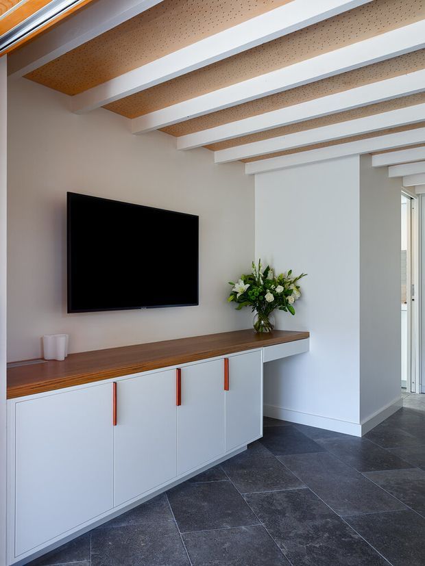 A bright contemporary living area with a wall-mounted television above a long built-in sideboard. The sideboard has white flat-panel cabinetry, a dark timber benchtop, and slim red-orange handles. The room features dark large-format floor tiles, pale walls, and a distinctive ceiling with exposed light timber acoustic panels between painted white beams. Glazed doors or windows open to the side, and a floral arrangement sits on the console.