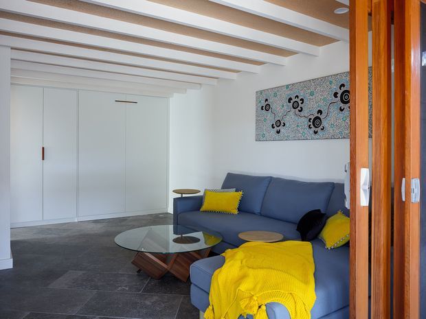 A bright contemporary living room viewed through open timber-framed doors. The room features a blue sectional sofa with vivid yellow cushions and throw, a transparent glass coffee table with sculptural wooden supports, dark large-format tiled flooring, white built-in storage, and a white ceiling with exposed timber beams. A patterned artwork adds a graphic accent above the sofa.