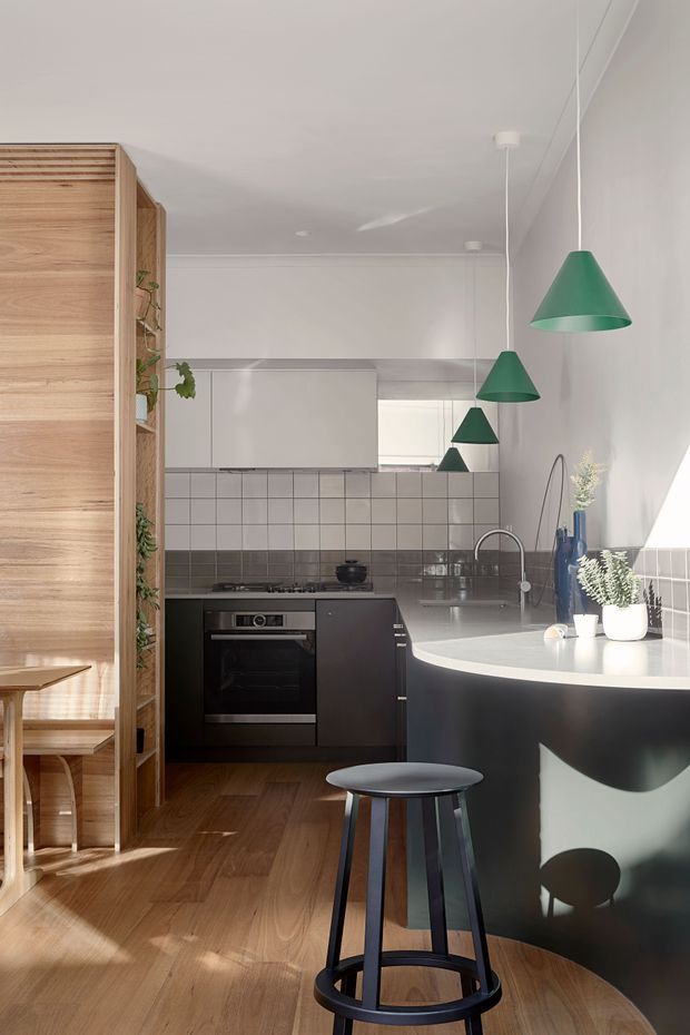 A compact contemporary kitchen with warm timber flooring and a timber slatted room divider, dark lower cabinetry, white upper cabinetry, and a tiled splashback in pale and dark grey tones. A curved island with a light benchtop dominates the foreground, paired with a black bar stool. Green pendant lights hang above the island, while a large mirror reflects the kitchen and pendants. Plants and simple accessories add a restrained natural accent.