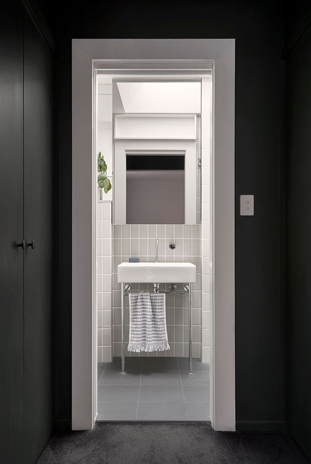 A compact bathroom viewed through a doorway, featuring a wall-mounted white porcelain basin with exposed chrome plumbing, a grey tiled floor, light grey square tiled walls, and a mirrored or dark reflective cabinet above. The bathroom is framed by dark charcoal walls and white trim, with a small plant and patterned hand towel adding subtle detail. A high-level angled skylight or highlight window brightens the space.