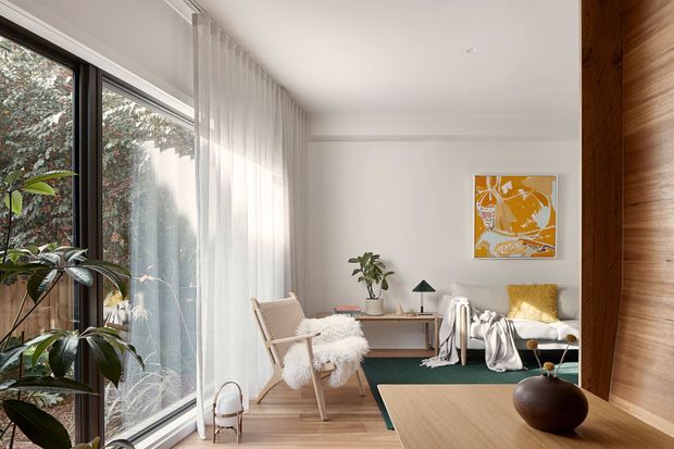 A bright, contemporary living room with full-height glazing along one side, sheer white curtains, pale timber flooring, and a warm timber feature wall or built-in joinery at right. The room includes a light lounge chair, sofa, small desk or console, indoor plants, a dark green rug, and a low timber coffee table with sculptural decor. An abstract orange artwork adds a vivid accent to the otherwise soft neutral palette.