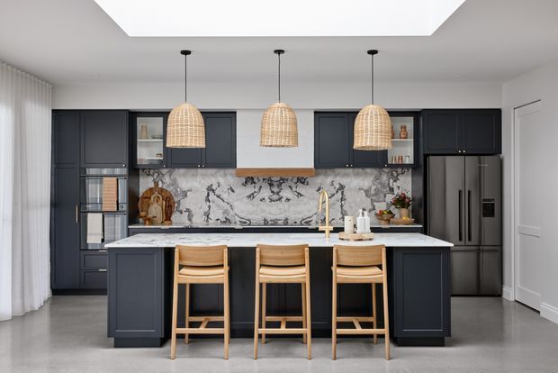 A refined contemporary kitchen viewed straight on, with a large island and three timber bar stools, dark navy-blue shaker-style cabinetry, a dramatic veined marble-look splashback, pale stone benchtops, integrated appliances, and three woven pendant lights. White walls, sheer curtains, and a large ceiling cut-out/skylight-like feature create a bright, polished setting.
