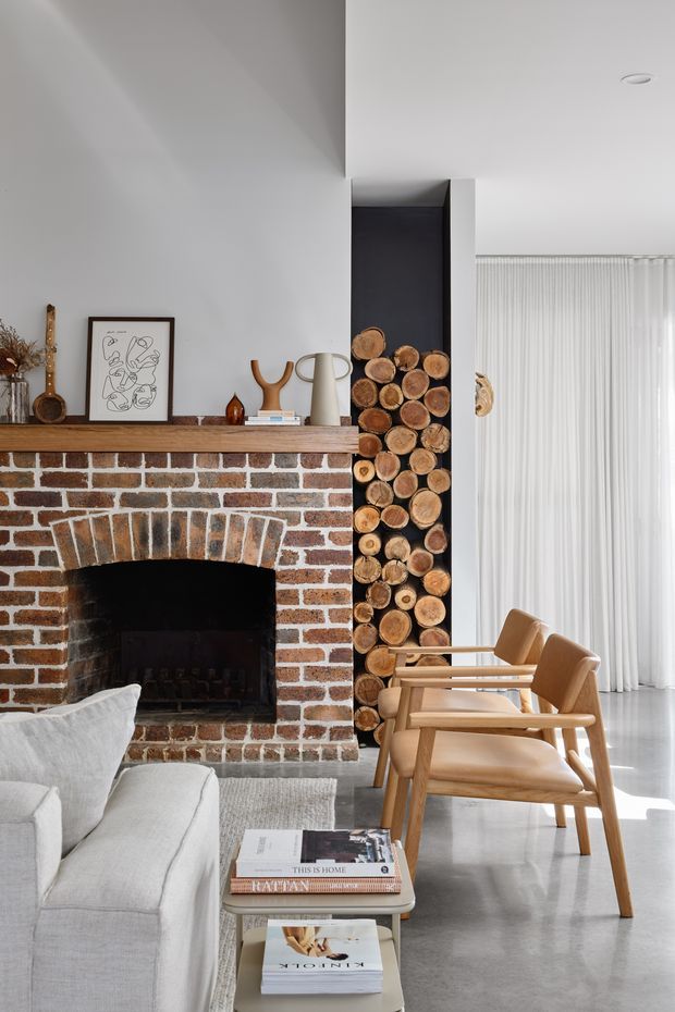 Bright contemporary living room featuring a red brick fireplace with a timber mantel, a recessed vertical firewood display, pale upholstered seating, timber-framed armchairs, a low coffee table, and full-height sheer curtains. The room combines polished concrete flooring, white walls, warm timber accents, and carefully styled decor.