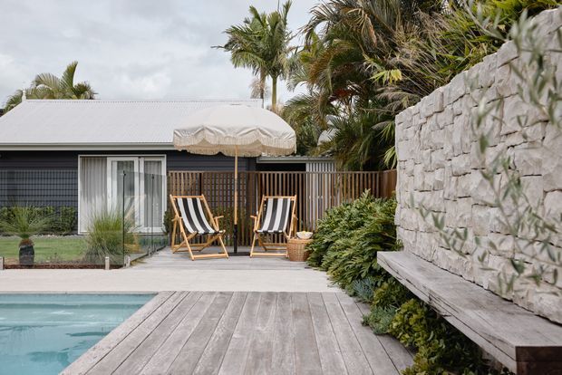 A stylish tropical outdoor pool area beside a low modern building, featuring a timber deck, pale stone feature wall with integrated bench seating, timber privacy screen, striped deck chairs, a large fringed umbrella, and lush palm planting. The scene has a relaxed resort-like atmosphere with muted natural materials and a cool grey palette.