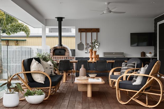 A bright, open-plan indoor-outdoor living and dining area with a timber-look floor, white vertical wall panelling, and extensive glazing opening toward a landscaped exterior. The room features rattan lounge chairs with dark cushions, a timber coffee table, a dining table with black chairs, a built-in brick pizza oven, a wall-mounted television, and warm natural styling. A ceiling fan and covered outdoor setting create a relaxed entertaining space.