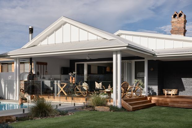 Exterior view of a single-storey residential home with a prominent gabled roof, white weatherboard-style cladding, covered outdoor entertaining veranda, timber decking and steps, and a small swimming pool in the foreground. The facade combines light painted cladding with dark horizontal wall sections, a brick chimney, glazed doors, and relaxed outdoor seating.