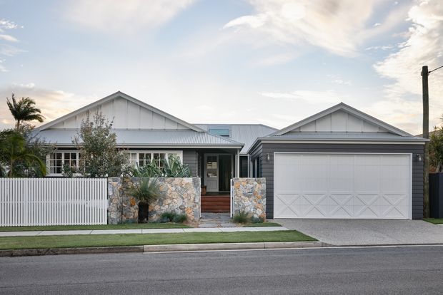 Street-facing view of a contemporary single-storey suburban house with two gabled roof forms, light grey metal-clad facades, a large integrated double garage, front garden, and a low stone-and-slatted fence. The entry is recessed between the main house and garage, with landscaped planting and a paved driveway.