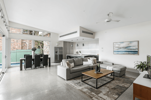 Bright open-plan living area with a contemporary kitchen and dining zone. The room features polished concrete flooring, white walls and ceiling, large glazed openings, a grey sectional sofa, timber-and-metal coffee table, compact dining setting, stainless-steel refrigerator, and minimalist white cabinetry. A ceiling fan and linear ventilation grilles are visible, with a courtyard and brick exterior seen through the glazing.