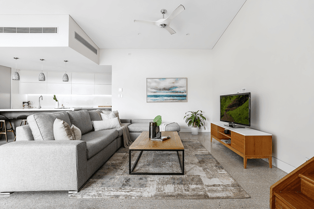 A bright, open-plan contemporary living room with a large light-grey sectional sofa, rectangular timber-and-metal coffee table, patterned area rug, timber television console, and wall-mounted TV. The adjoining white kitchen includes a peninsula with stools, pendant lights, white cabinetry, and stainless-steel appliances. A stair with timber treads is visible at the right edge, along with a ceiling fan, indoor plants, and a coastal-toned artwork.
