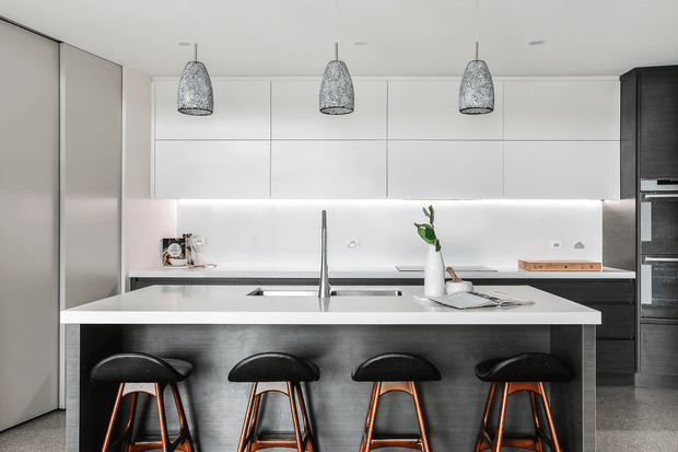 Contemporary kitchen with a large central island, four black bar stools, white flat-panel cabinetry, dark timber-toned tall cabinetry, integrated appliances, and three textured pendant lights. The island includes a sink and faucet, with a light neutral palette and subtle timber and copper accents.