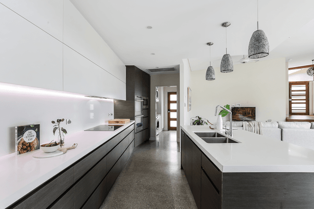 Bright contemporary open-plan kitchen with a long dark timber island and matching cabinetry, white engineered-looking benchtops, integrated appliances, pendant lights, and a small living area visible beyond. The space features polished concrete flooring, white upper cabinetry, and timber-framed glazing at the rear.