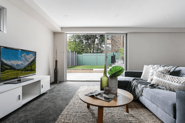 Bright contemporary living room with a grey sectional sofa, textured area rug, round timber coffee table, white entertainment unit, and television. Large sliding glass doors open to a small fenced outdoor area, bringing in natural light. The room has neutral finishes, recessed ceiling lights, and minimal decorative styling.