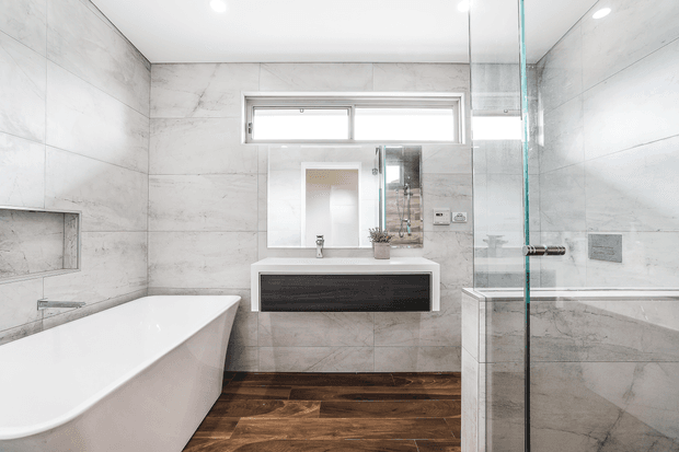 Contemporary bathroom with a freestanding white bathtub, wall-mounted dark timber vanity, large-format pale grey stone-look wall tiles, frameless glass shower enclosure, recessed wall niche, and dark timber-look flooring. A wide horizontal window and mirrored vanity area bring natural light into the clean, refined space.