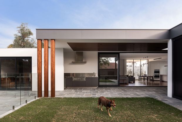 Exterior view of a contemporary single-storey residence with a broad flat roof, deep covered outdoor area, white rendered walls, dark-framed glazing, and prominent vertical timber posts. The open terrace is paved in large grey tiles and looks into a modern kitchen and dining area, with a small lawn in the foreground.