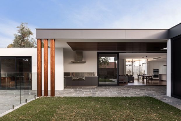 Contemporary single-storey residence viewed from the rear courtyard, with a broad flat-roofed covered opening connecting an outdoor paved area to an open-plan kitchen, living and dining zone. The facade combines smooth white rendered walls, dark-framed glazing, charcoal cabinetry and a prominent vertical timber-clad feature beside the kitchen. A lawn and glass balustrade occupy the foreground, while the interior shows a kitchen, dining table and living area beyond.