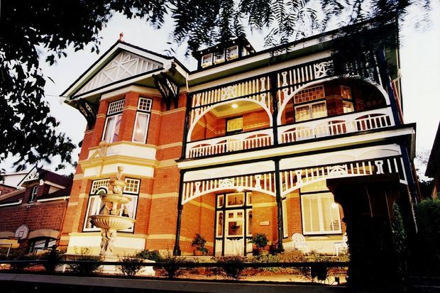 Exterior view of a large ornate red-brick multi-storey residence with prominent white-painted timber verandahs and decorative balustrades, projecting bay windows, gabled roof sections, and classical detailing. Mature trees frame the facade and a fountain sits in the front garden.