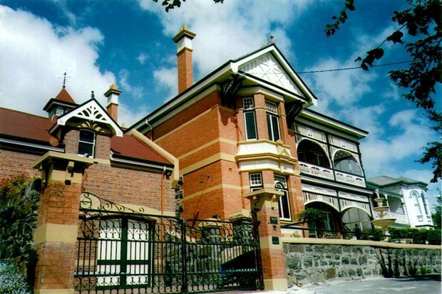 Exterior view of a large historic brick residence with ornate Victorian detailing, steep gabled roofs, tall chimneys, decorative timber gable trim, projecting bay windows, elaborate verandahs and balconies, wrought-iron gates, and a stone boundary wall. The image has a vivid, high-contrast photographic appearance and shows a substantial multi-level heritage-style home.