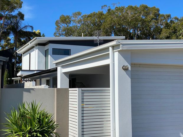 Exterior view of a contemporary two-storey residential home with crisp white rendered walls, a low-pitched roof, horizontal louvre-style garage door, sheltered entry, and a small landscaped frontage. Dark-framed horizontal windows and clean geometric lines create a modern facade.