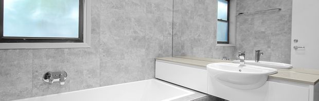 A contemporary bathroom with a white freestanding-style bathtub, wall-mounted bath fixture, floating white vanity with integrated basin, and large frameless mirror. Grey stone-look wall tiles and matching grey surfaces create a clean, cool-toned scheme, with black or dark metal tapware and narrow windows providing natural light.
