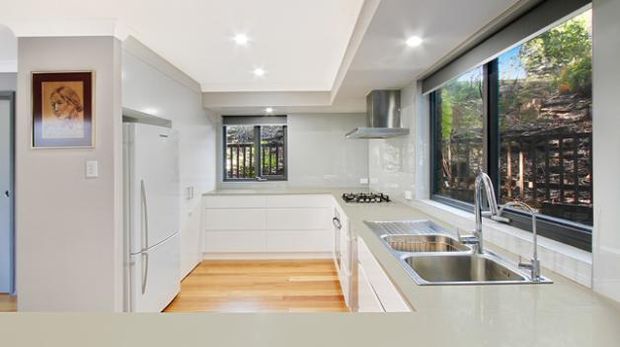 Bright contemporary galley kitchen with white flat-panel cabinetry, a long pale benchtop, stainless-steel double sink and gooseneck tap, integrated cooktop and rangehood, large refrigerator, timber flooring, and a wide window overlooking greenery. Recessed ceiling lights and clean-lined finishes create an airy, practical interior.