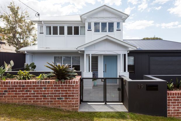 Street-facing exterior of a two-storey white weatherboard home with a prominent gabled entry porch, louvred windows, light blue front door, dark garage, and a low red-brick boundary wall with landscaped planting.