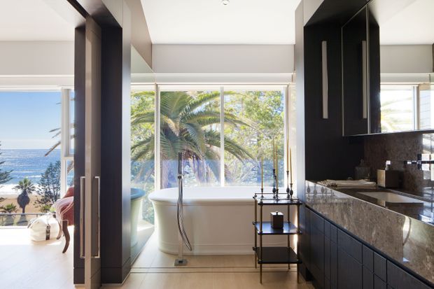 A luxurious open-plan bathroom with a freestanding bathtub positioned beside expansive floor-to-ceiling glazing overlooking a coastal scene. Dark full-height cabinetry and a dark stone vanity contrast with pale flooring and walls, while a bedroom or adjacent living space is visible through the opening at left.