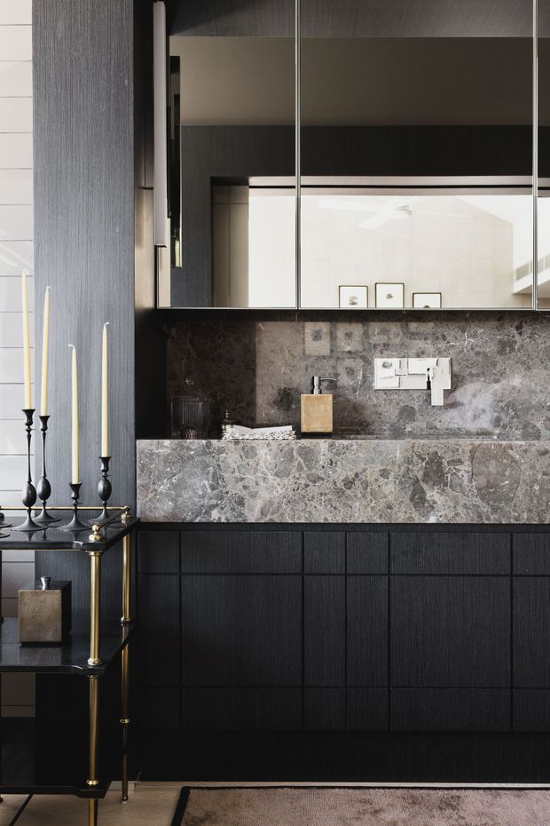 A sophisticated dark-toned bathroom vanity featuring a large grey marble benchtop and matching splashback, black fluted cabinetry, mirrored wall cabinets, and polished chrome tapware. A black and brass console with tall candlesticks sits to the left, while framed artwork is reflected in the mirrors. The overall composition is luxurious, moody, and highly refined.