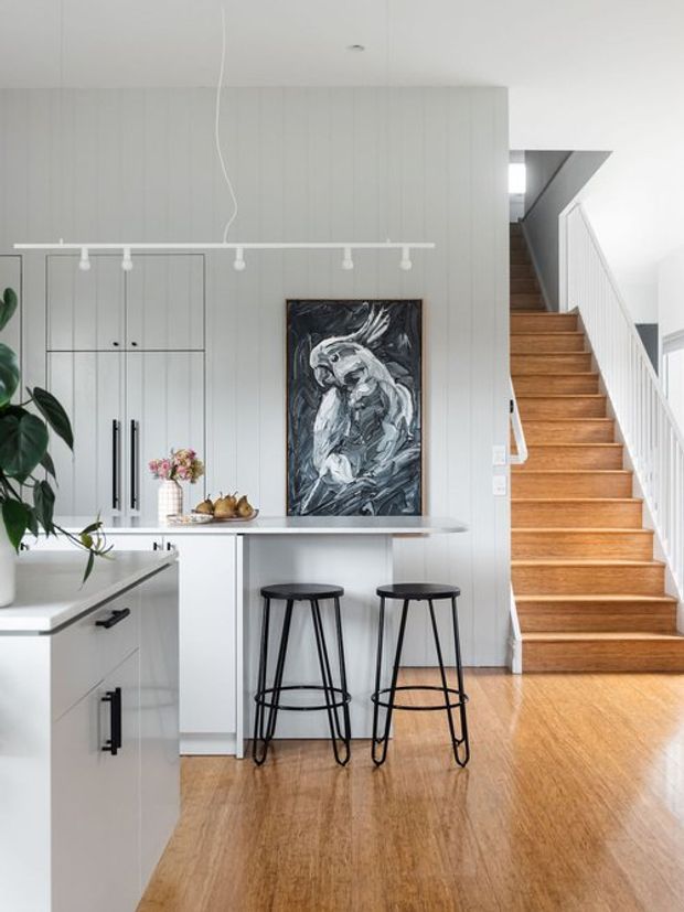 Bright, compact open-plan kitchen interior with white vertical wall panelling, integrated white cabinetry, a pale island with a light benchtop, and two black metal bar stools. A large monochrome artwork hangs above the island, while a timber staircase with white balustrade rises along the right side. Warm timber flooring and abundant natural light create a clean, inviting space.