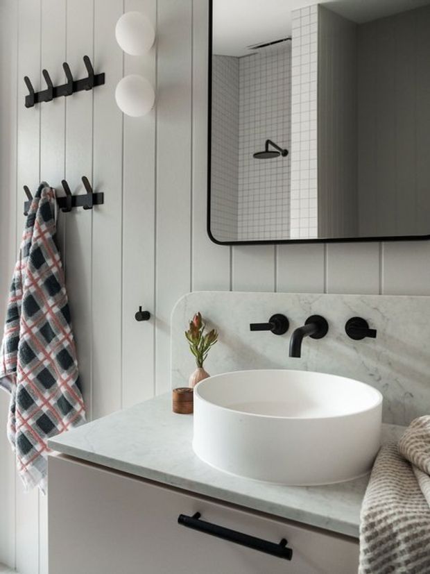 A refined bathroom vanity with a white vessel basin, marble or stone benchtop and matching splashback, matte black wall-mounted tapware, and a large rounded-rectangle mirror. Vertical white wall panelling, black hooks, soft towels, and simple spherical wall lights create a clean contemporary look with subtle traditional detailing.