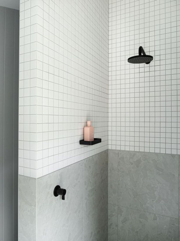 A minimalist shower enclosure with white square wall tiles above grey stone-look lower wall tiles. Matte black shower hardware and a small black shelf with a blush-pink bottle create restrained contrast. The image is tightly framed on the tiled shower area, with no visible vanity or bedroom connection.