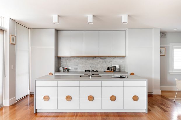 Bright contemporary kitchen centered on a large white island with five round timber pulls. The room features extensive flat-panel white cabinetry, a pale stone-look benchtop, a grey-white exposed brick splashback, polished timber flooring, and simple white ceiling-mounted linear lights. A dining area is partially visible to the right.