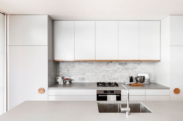 Bright, streamlined contemporary kitchen with extensive white flat-panel cabinetry, a grey stone-look benchtop, white brick-pattern splashback, integrated oven and gas cooktop, and a large island with undermount sink and gooseneck tap. The space has minimal detailing and a clean neutral palette.