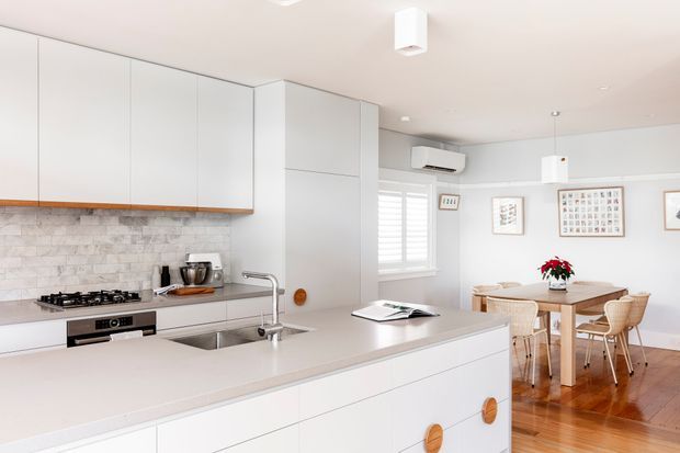 Bright open-plan kitchen and dining area with white flat-panel cabinetry, a pale stone island benchtop, stainless-steel sink and tap, gas cooktop, and a light textured stone splashback. The adjacent dining area has a timber table and woven chairs, with polished timber flooring, plantation shutters, framed artwork, and simple pendant lighting.