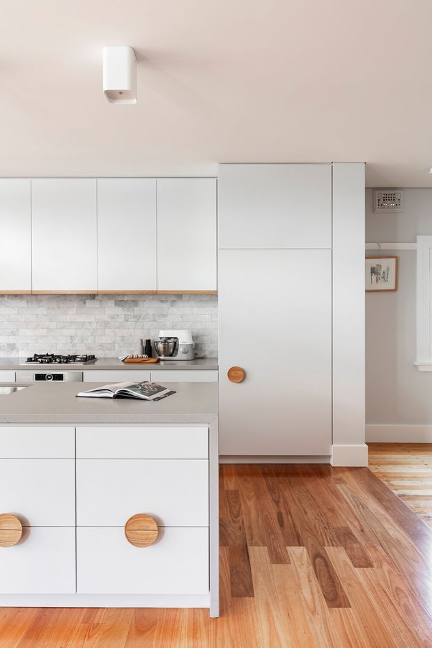 Bright, compact contemporary kitchen with white flat-panel cabinetry, a pale stone island benchtop, and integrated tall storage. A light grey-white brick-style tiled splashback runs behind the cooktop and benchtop appliances. Warm timber flooring contrasts with the soft neutral walls and cabinetry, while round timber pulls add a simple natural accent. A small adjoining area is visible to the right.