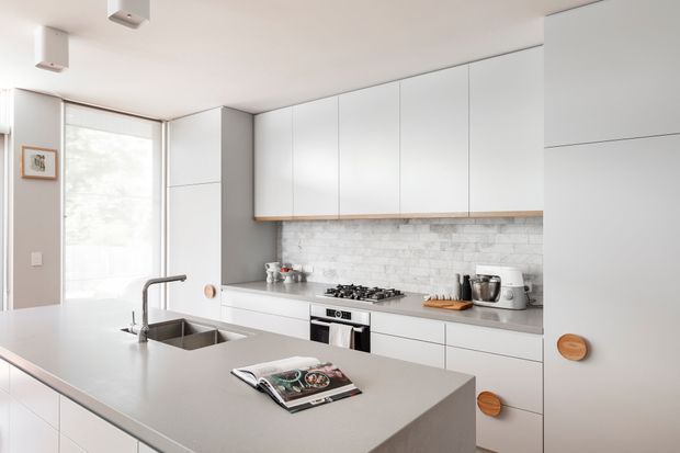 A bright, streamlined contemporary kitchen with a large island, integrated double sink and gooseneck tap in the foreground. White flat-panel cabinetry runs across the back wall, complemented by a pale grey benchtop and a white-grey textured stone-tile splashback. A gas cooktop, built-in oven, small appliances and timber shelf detail are visible, with full-height glazing bringing in abundant natural light.