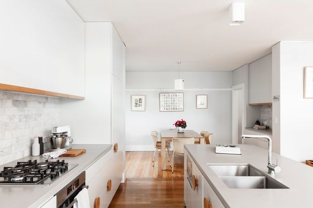 A bright, compact open-plan kitchen viewed toward an adjoining dining area. White flat-panel cabinetry lines both sides, with a pale stone-look tiled splashback, light grey benchtops, stainless-steel double sink and tapware, gas cooktop, and integrated appliances. Warm timber flooring leads to a small timber dining table and chairs beneath simple pendant lighting. The room has a clean, restrained contemporary feel with soft grey walls and white ceilings.