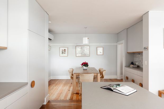 Bright, compact open-plan kitchen and dining area viewed from the kitchen. White built-in cabinetry frames the space, with a grey benchtop in the foreground and a recessed kitchen workspace along the right wall. A timber dining table and woven chairs sit on light timber flooring beneath a simple pendant light. Pale grey walls, framed artwork, and restrained detailing create a clean, calm interior.