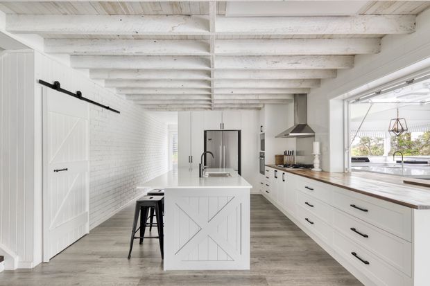 Bright, elongated farmhouse-style kitchen with white cabinetry, a central island, stainless-steel appliances, timber benchtops, and exposed white-painted ceiling beams. A white barn-style sliding door and white brick wall reinforce the rustic architectural character, while large glazing brings in abundant natural light.