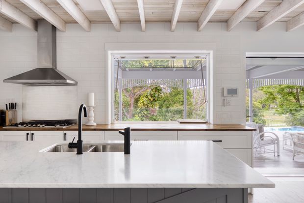 Bright, spacious kitchen with a large white stone island, integrated sink and black fixtures, white cabinetry, timber benchtop accents, and a stainless steel rangehood. White-painted exposed ceiling beams and light masonry-style wall tiling add architectural character. Full-height glazing and an open glazed door connect the kitchen to a covered outdoor area and pool beyond.