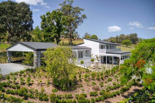 A white two-storey rural residence with a broad covered veranda and attached open-sided carport, set within landscaped gardens on a gently sloping green site. The home has a dark pitched metal roof, light exterior cladding, stone-clad carport columns, and traditional veranda detailing. Bright daylight and mature trees surround the property.