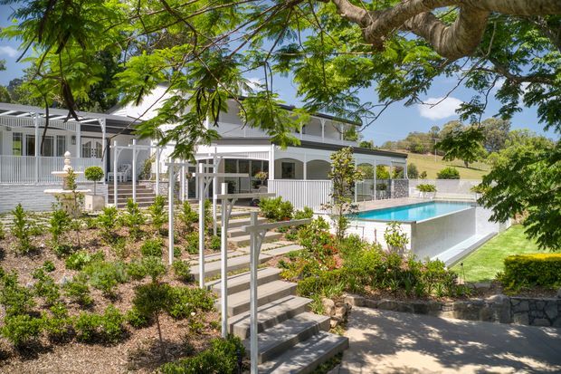 Exterior view of a large light-coloured residence set within landscaped gardens, featuring a broad verandah or covered outdoor living area, white railings and columns, a pitched metal roof, and a raised rectangular swimming pool. Stone retaining walls, planted garden beds, steps, and mature trees surround the house.