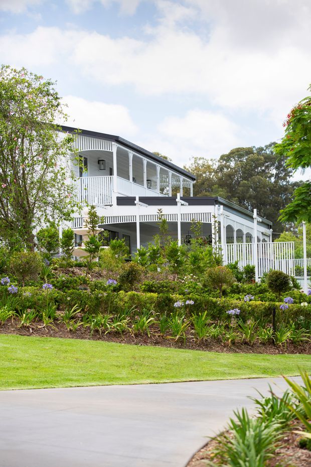 Exterior view of a large white multi-level residence with wraparound verandas, decorative railings, covered outdoor living areas, and arched openings. The home is set within a carefully landscaped garden with flowering plants, shrubs, lawn, and a paved driveway. The design has a traditional Australian veranda-house character with a dark pitched roof and extensive white detailing.